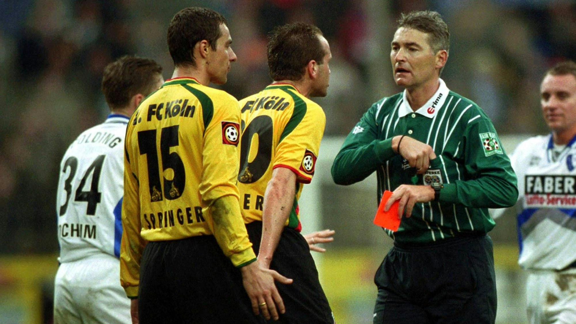 Zwei Spieler des 1. FC Köln in gelben Trikots diskutieren mit dem Schiedsrichter, der eine rote Karte in der Hand hält.