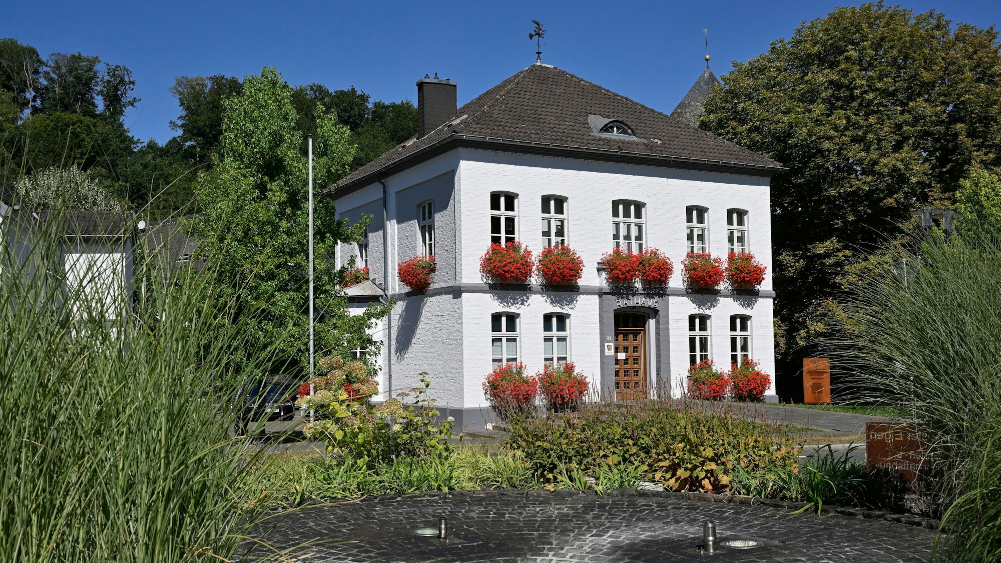 Das Rathaus in Odenthal mit roten Geranien vor den Fenstern. Vor dem Gebäude befindet sich ein Springbrunnen.