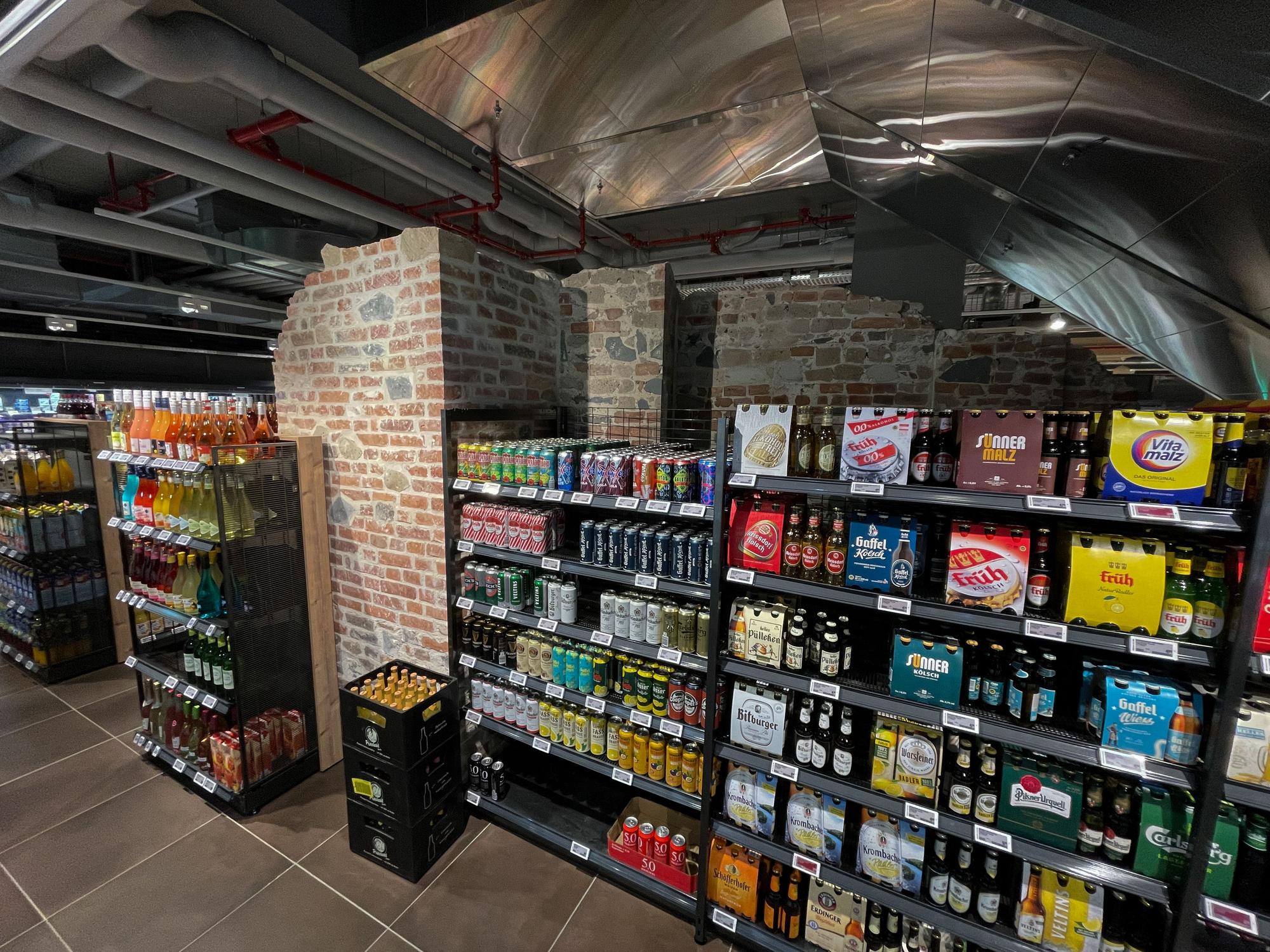 Ein Getränkeregal im Rewe, dazwischen ein Stück Stadtmauer.