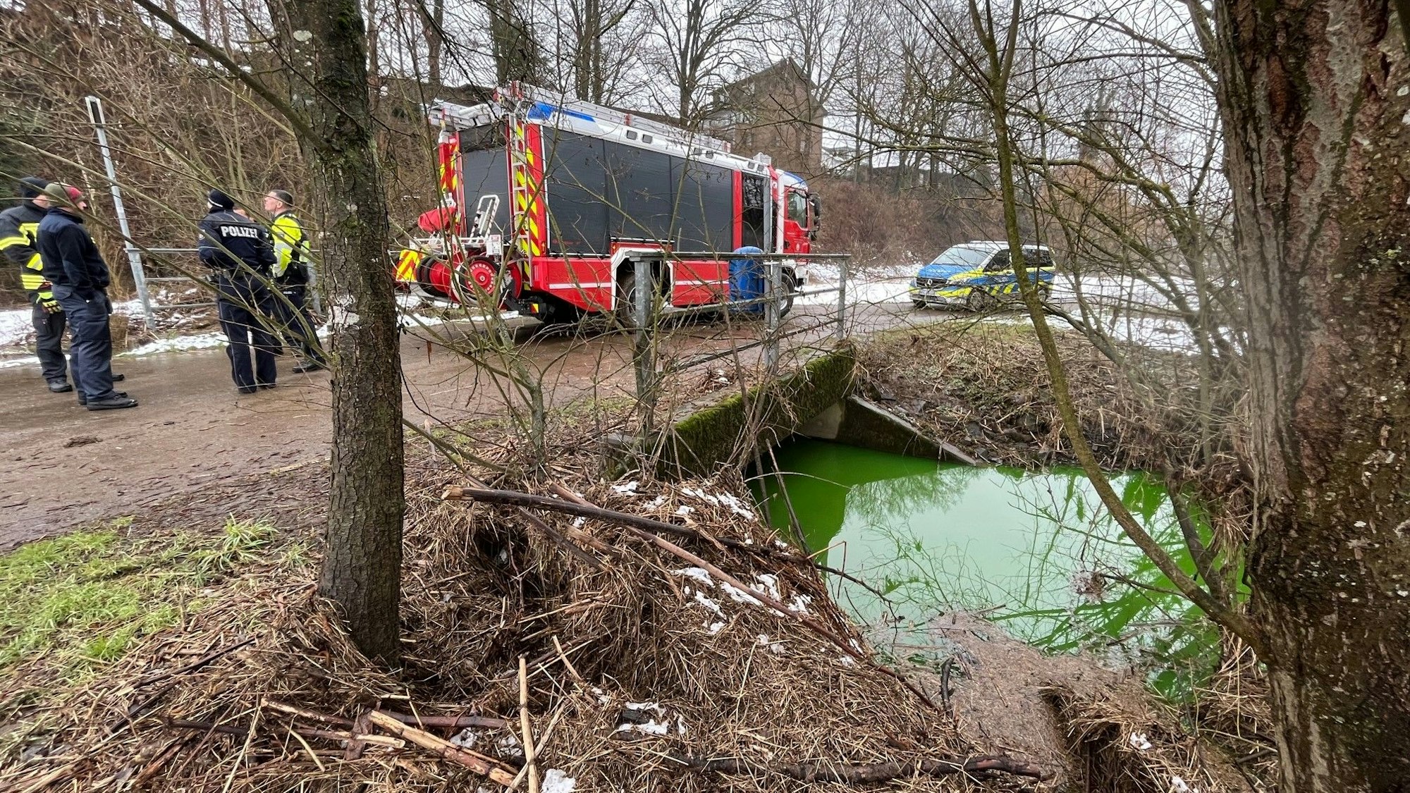 Feuerwehrleute und Polizisten stehen auf einer kleinen Brücke über den grünen Bach, ein Löschfahrzeug und ein Polizeiwagen stehen auf dem schlammigen Weg.