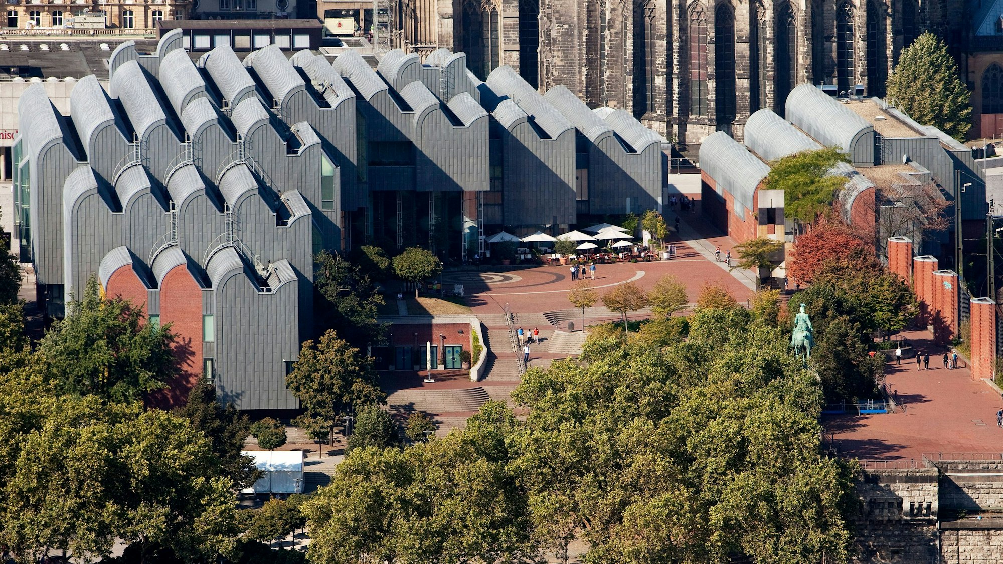 Blick von oben auf den Gebäudekomplex Kölner Philharmonie und Museum Ludwig mit seinem charakteristischen wellenförmigem Dach. Im Hintergrund sind die Mauern des Kölner Doms zu sehen.