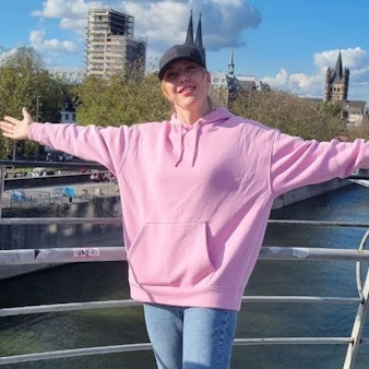 Eine Frau in einem rosa Sweater und ausgebreiteten Armen vor dem Rheinpanorama