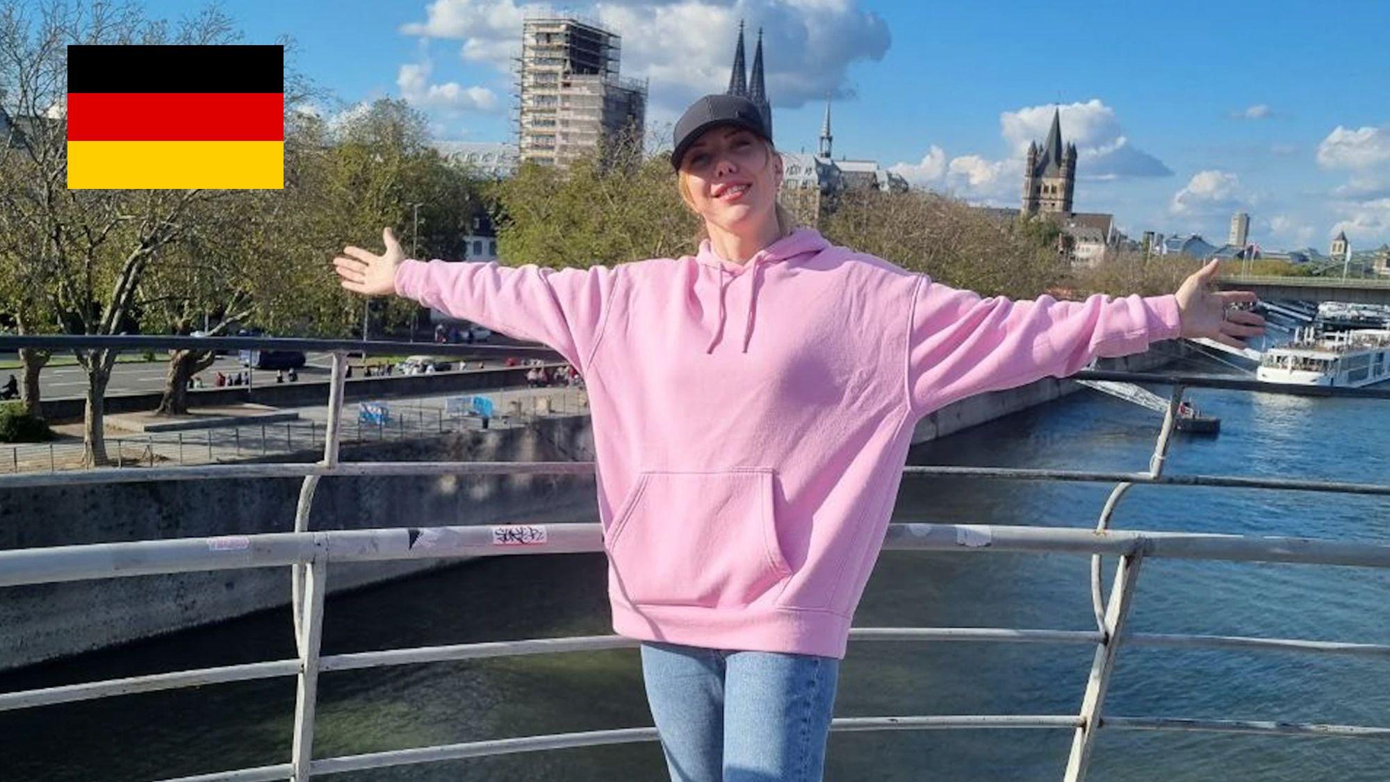 Eine Frau in einem rosa Sweater und ausgebreiteten Armen vor dem Rheinpanorama