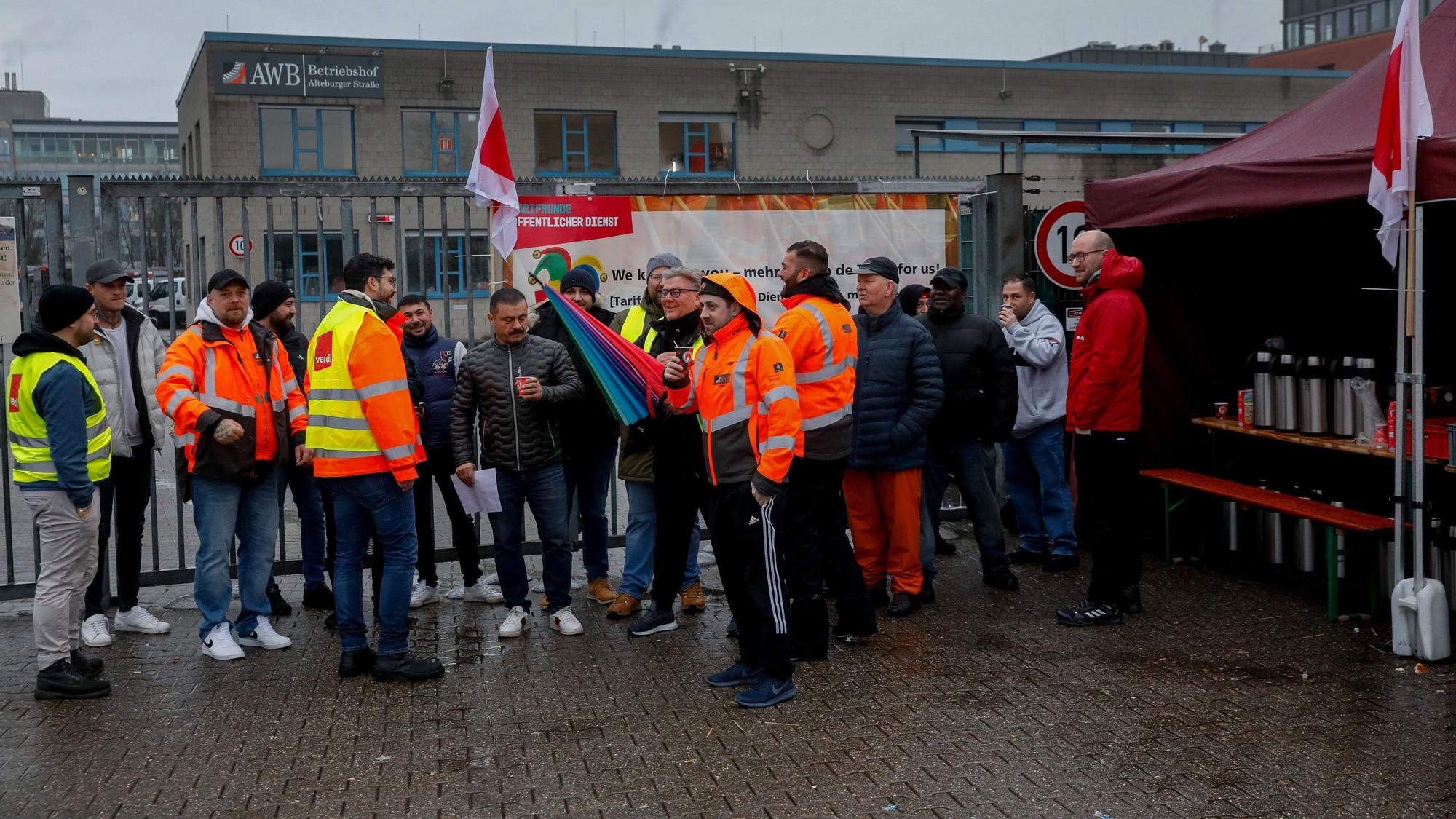 Männer in orangenen AWB-Jacken stehen vor dem Betriebshof der AWB. Manche tragen zusätzlich Warnwesten von Verdi.
