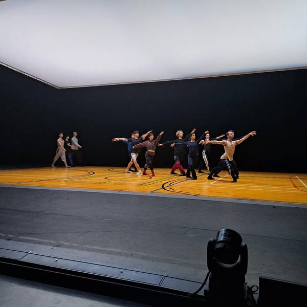 Eine Gruppe an Tänzerinnen und Tänzern steht in der gleichen Ballettposition in der Mitte der Bühne. Sie haben ausgestreckte Arme und Beine. Eine andere kleinere Gruppe geht im Hintergrund im Gleichschritt.