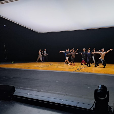 Eine Gruppe an Tänzerinnen und Tänzern steht in der gleichen Ballettposition in der Mitte der Bühne. Sie haben ausgestreckte Arme und Beine. Eine andere kleinere Gruppe geht im Hintergrund im Gleichschritt.