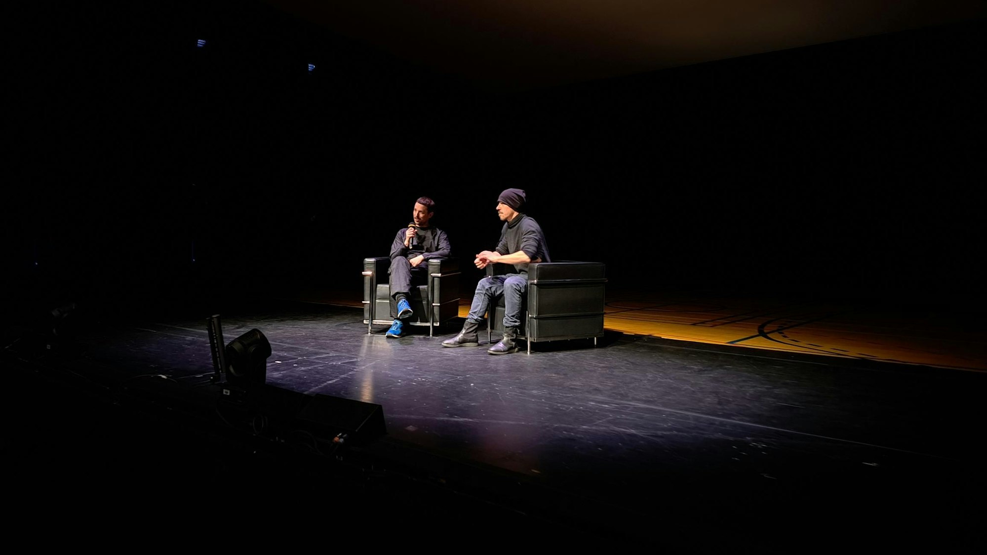 Dramaturg Tobias Staab (links) und Choreograf Richard Siegal sitzen auf Sesseln vor der Bühne im Depot 1 des Schauspiels Köln. Nur ein dunkler Lichtkegel beleuchtet sie. Staab hat ein Mikrofon in der Hand, beide gucken ins Publikum.