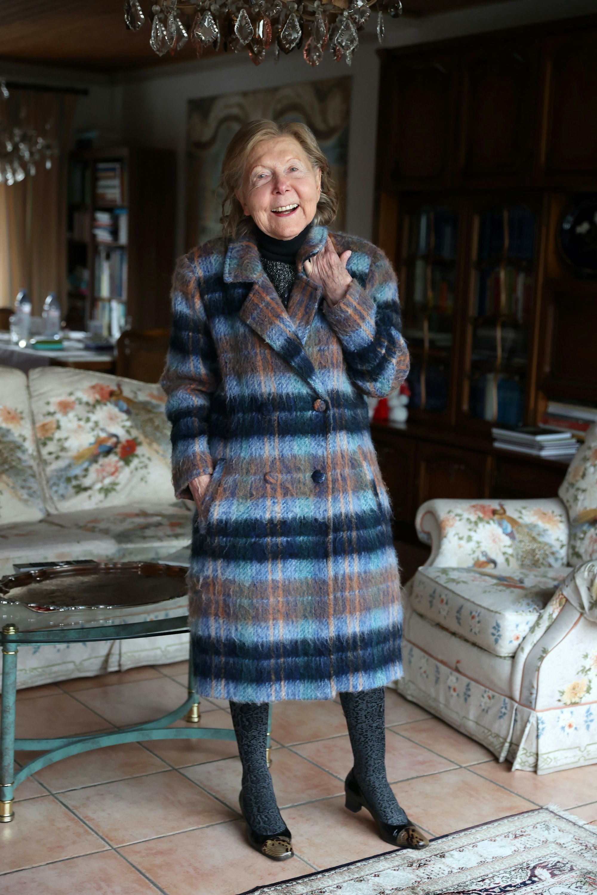 Renate Springer steht in ihrem Wohnzimmer und trägt den Mantel.