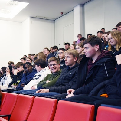 Eine Frau steht mit Mikrofon in der Hand vor mehreren Sitzreihen mit Schülern und spricht diese an.