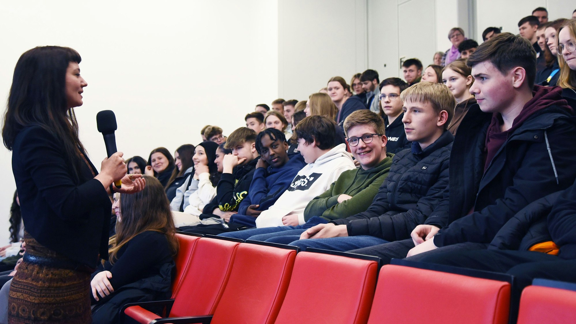 Eine Frau steht mit Mikrofon in der Hand vor mehreren Sitzreihen mit Schülern und spricht diese an.