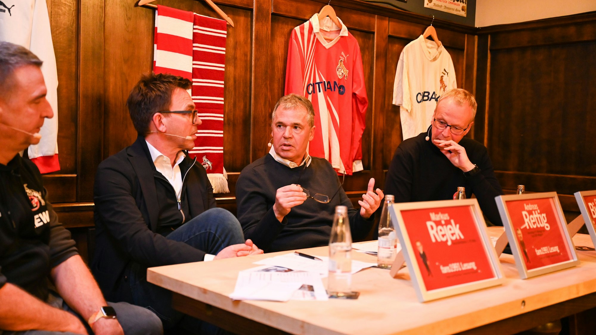 Fußballfunktionäre sitzen an einem Tisch und sind in eine Diskussion vertieft. Auf dem Tisch stehen Namensschilder, der Hintergrund ist mit Fanartikeln dekoriert.