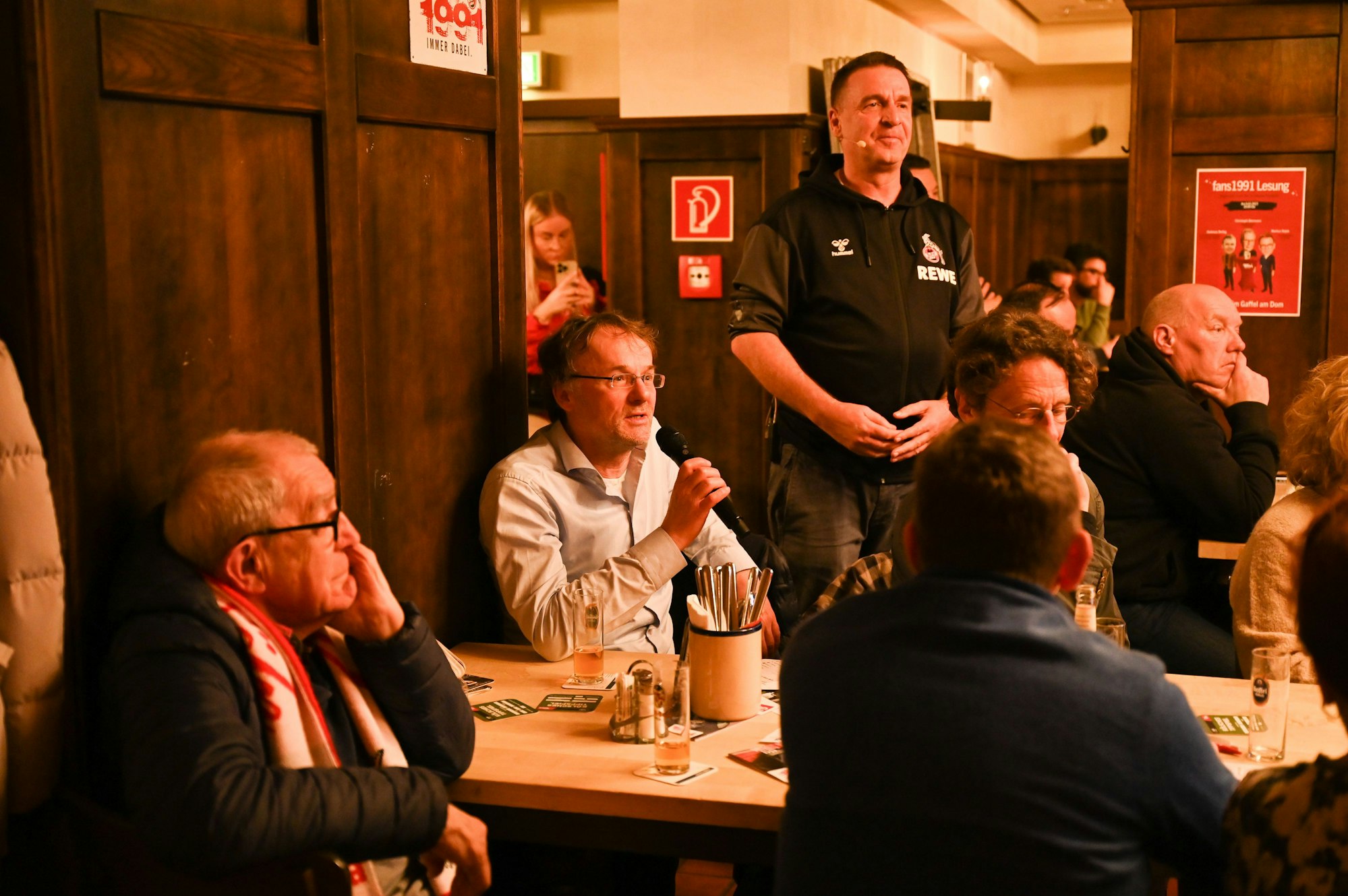 Ein Mann sitzt an einem Tisch und hält ein Mikrofon in der Hand. Die Umsitzenden hören interessiert zu.