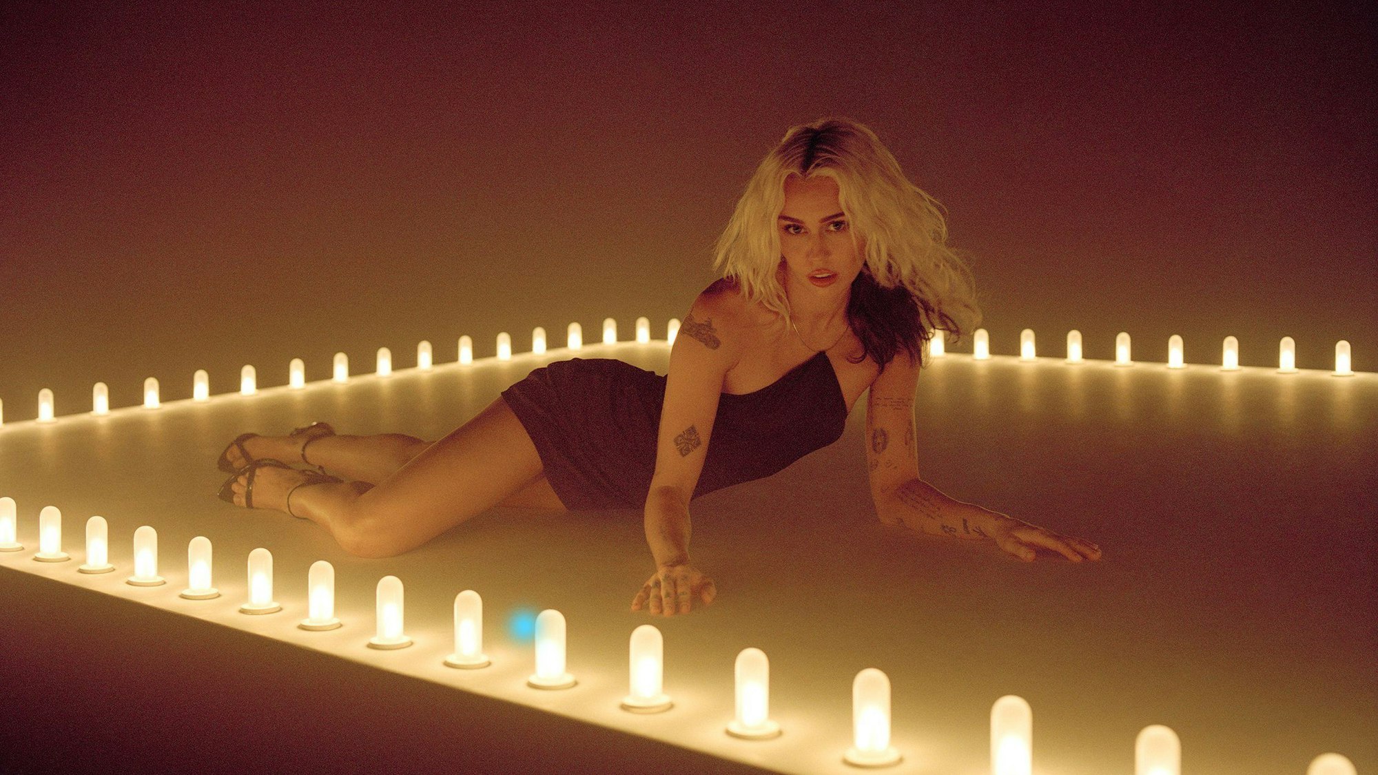 Miley Cyrus liegt zwischen Glühbirnen auf dem Boden, stützt sich auf ihren Armen ab und schaut lüstern in die Kamera. Es ist eine Szene aus dem Musikvideo zu ihrer neuen Single „River“.