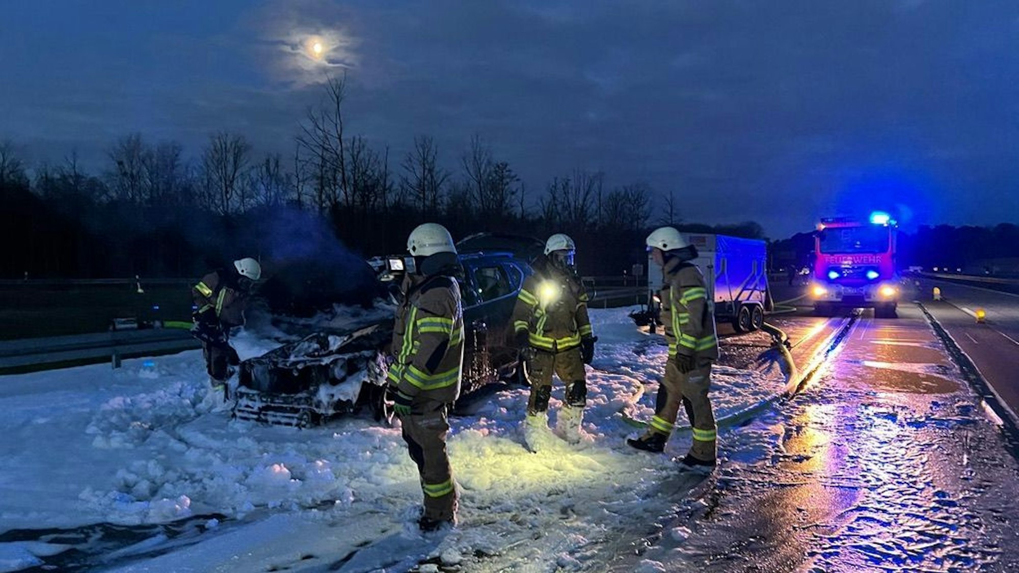 Feuerwehrleute stehen im Löschschaum um einen ausgebrannten Pkw herum. Im Hintergrund ist ein Löschfahrzeug zu sehen.