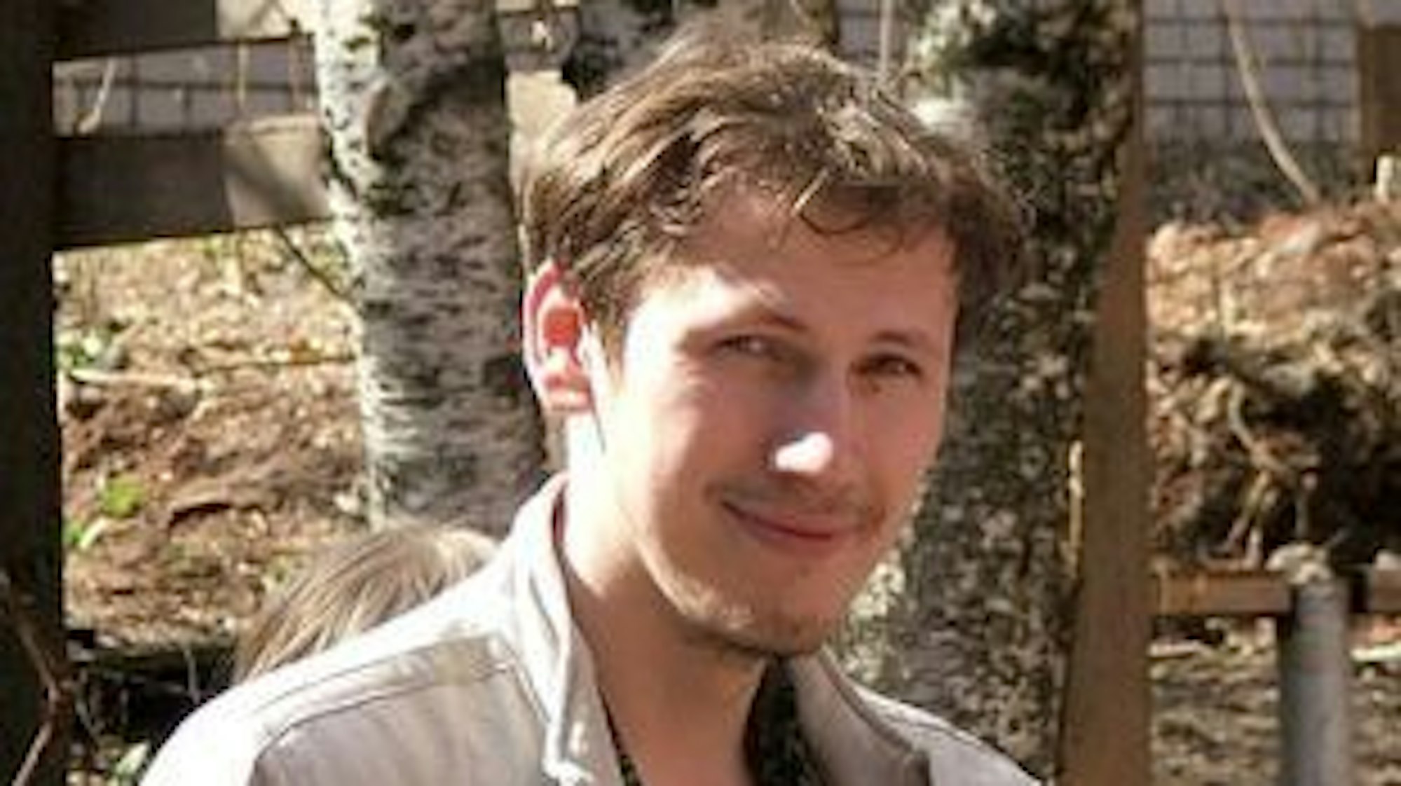 Igor Turashev steht in einem Park und guckt in die Kamera. Er hat dunkelblonde Haare und trägt einen weißen Mantel