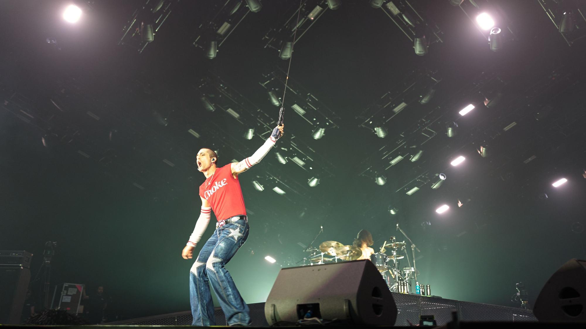 Måneskin-Frontman Damiano David steht auf der Bühne der Lanxess-Arena. Er hält das von der Decke hängende Mikrofon und schreit ins Publikum. Hinter im ist Schlagzeiger Ethan Torchio.