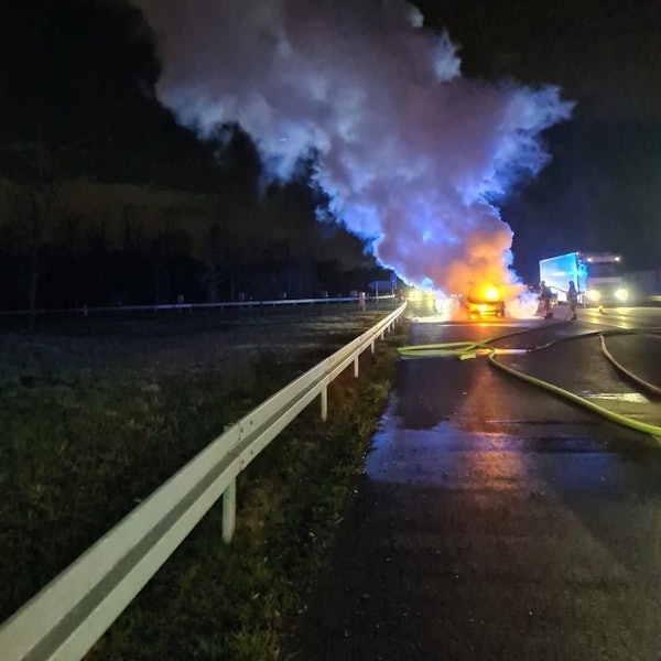 Feuerwehrleute löschen ein brennendes Auto auf dem Seitenstreifen der Autobahn 4. Ein Lkw fährt an der Einsatzstelle vorbei.
