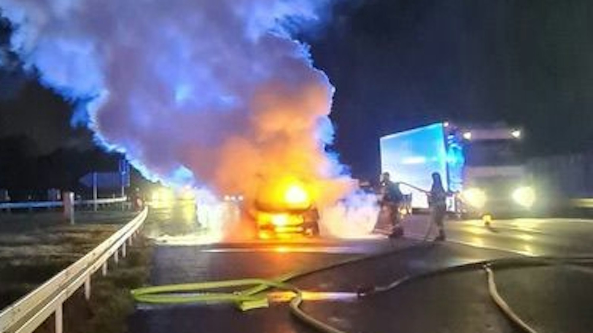 Feuerwehrleute löschen ein brennendes Auto auf dem Seitenstreifen der Autobahn 4. Ein Lkw fährt an der Einsatzstelle vorbei.