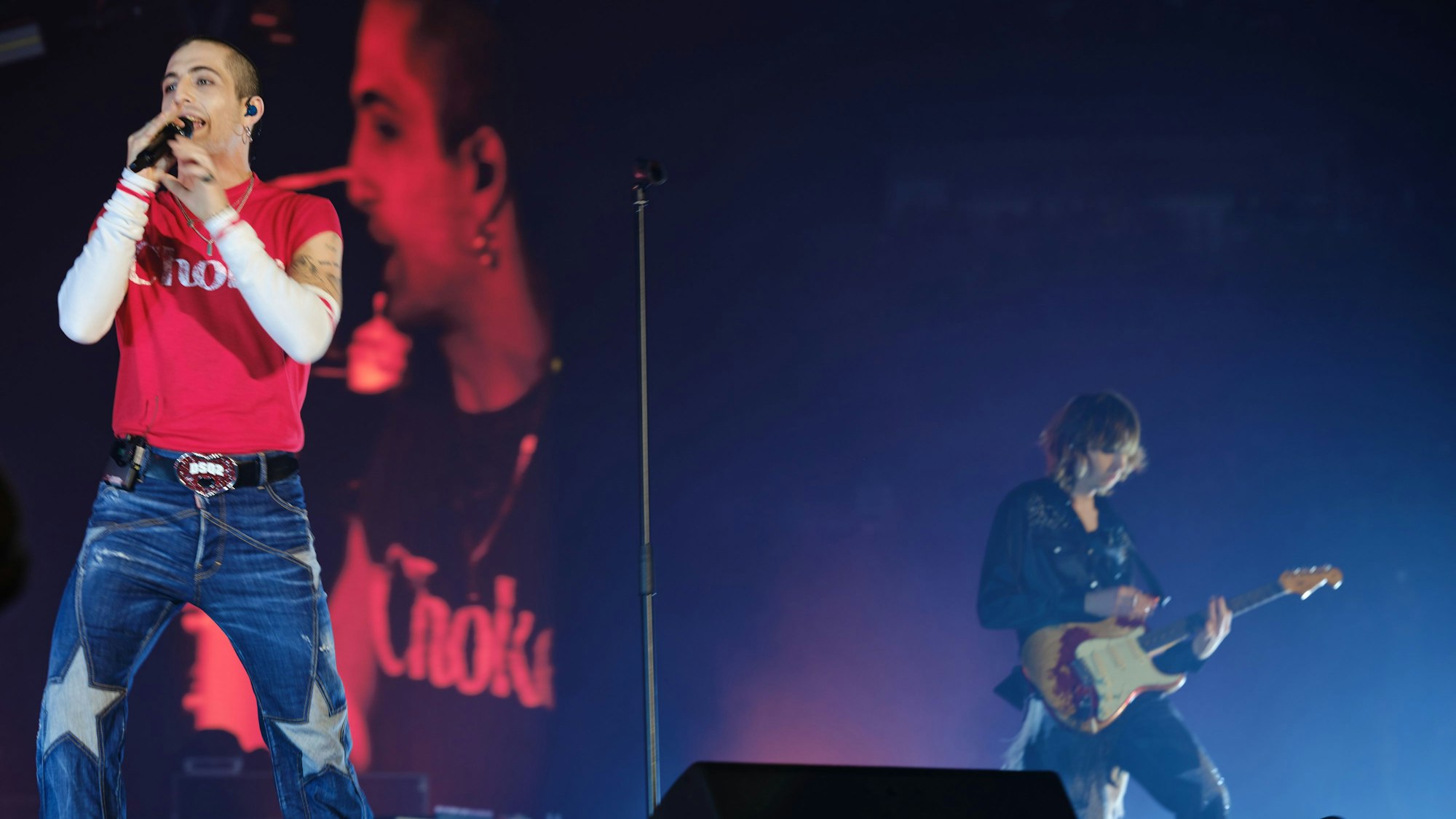 Im Vordergrund singt Damiano David ins Mikrofon und gestikuliert mit seiner anderen Hand. Im Hintergrund spielt Thomas Raggi E-Gitarre.