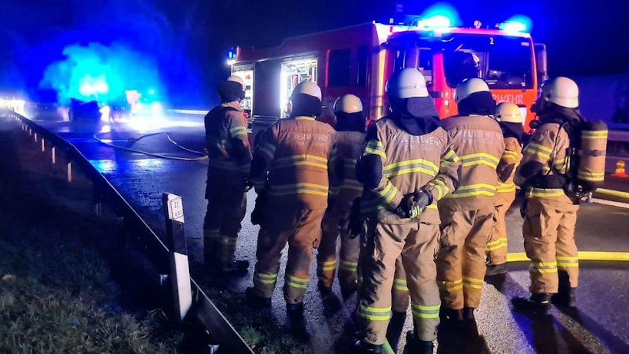 Feuerwehrleute stehen, teilweise mit Atemschutzausrüstung, auf dem Seitenstreifen der Autobahn. Im Hintergrund sind Einsatzfahrzeuge und ein brennender Pkw zu sehen.
