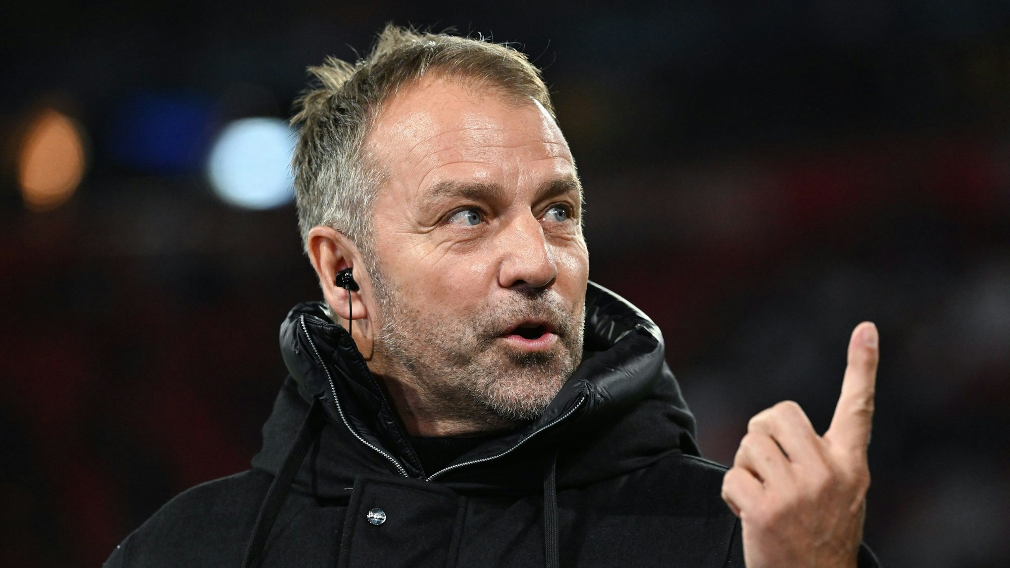 Bundestrainer Hansi Flick steht in einer schwarzen Jacke am Spielfeldrand, er hebt den Zeigefinger.