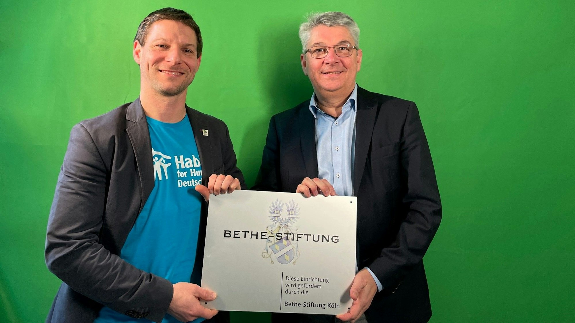 Der Vorstand der Organisation Habitat for Humanity, Gereon Fischer (links), und Lutz Urbach von der Bethe-Stiftung halten ein Schild der Bethe-Stiftung in Händen, das von der Stiftung geförderte Projekte kennzeichnet.