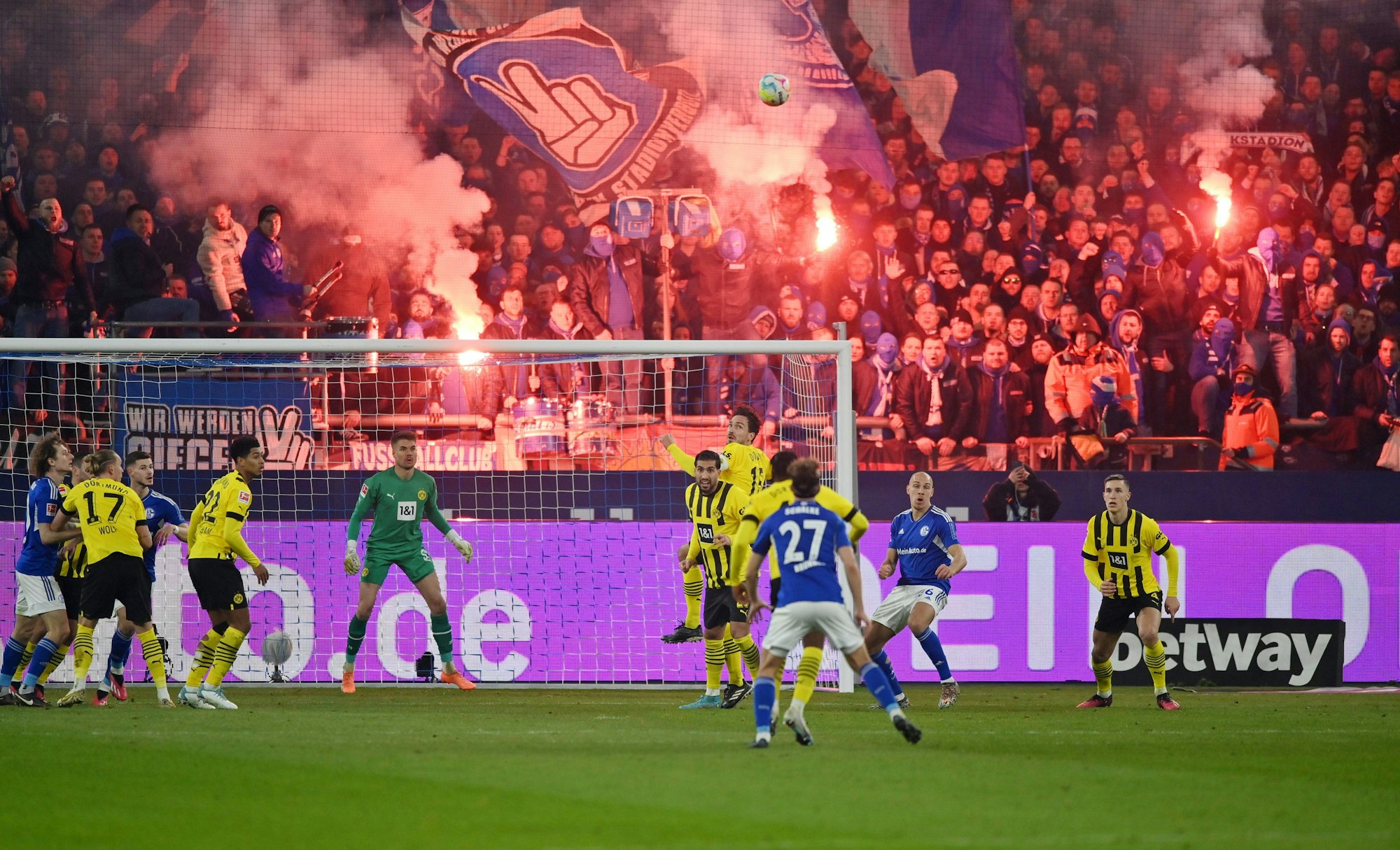 Spieler von Schalke und Dortmund kämpfen um den Ball, während im Hintergrund Fans bengalische Feuer abbrennen.