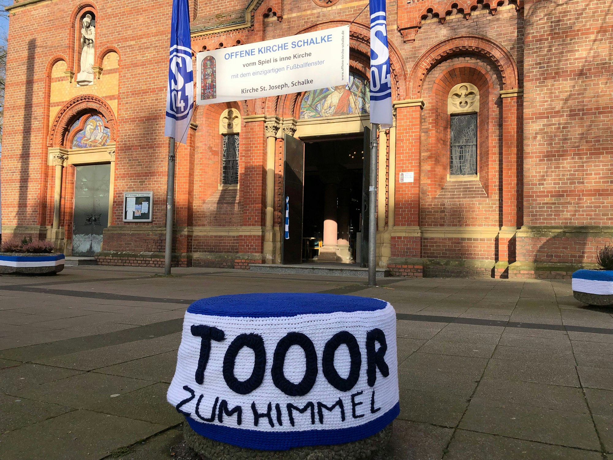 Ein gestrickter Überzieher in Blau und Weiß über einem Betonpoller. Darauf steht: Tooor zum Himmel. Im Hintergrund ist das Kirchenportal zu sehen, das von zwei Schalke-Fahnen flankiert wird.