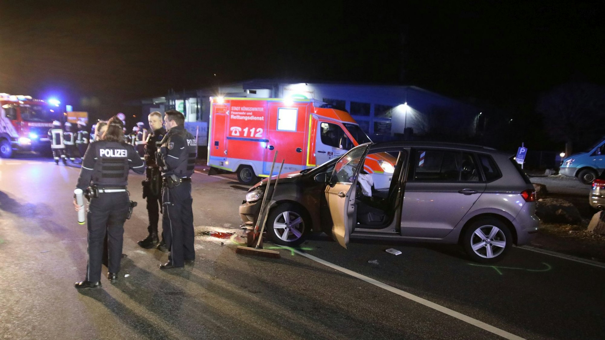 Ein verunfalltes Auto steht auf einer Straße, die Tür ist geöffnet, die Airbags wurden ausgelöst. Daneben stehen mehrere Polizisten und ein Rettungswagen. Es ist dunkel.