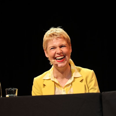 Cordula Stratmann sitzt in einem gelben Anzug auf der Bühne des Sartory-Saals.