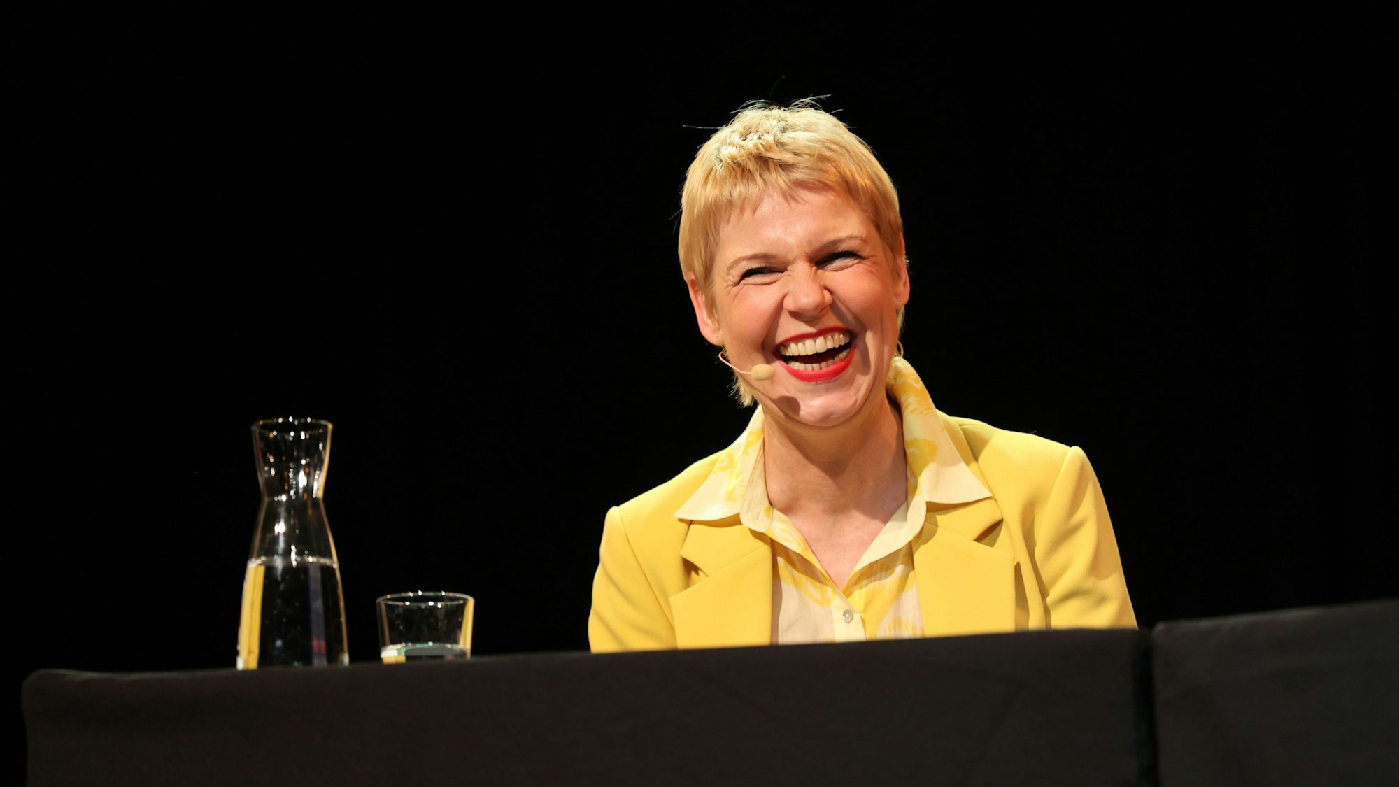 Cordula Stratmann sitzt in einem gelben Anzug auf der Bühne des Sartory-Saals.