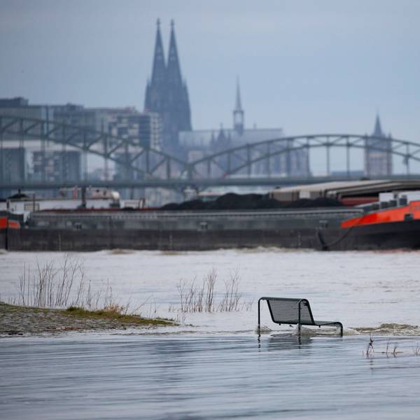 Binnenschiffe fahren auf dem Rhein vor der Kulisse des Kölner Doms, im Vordergrund steht eine vom Wasser umspülte Sitzbank.