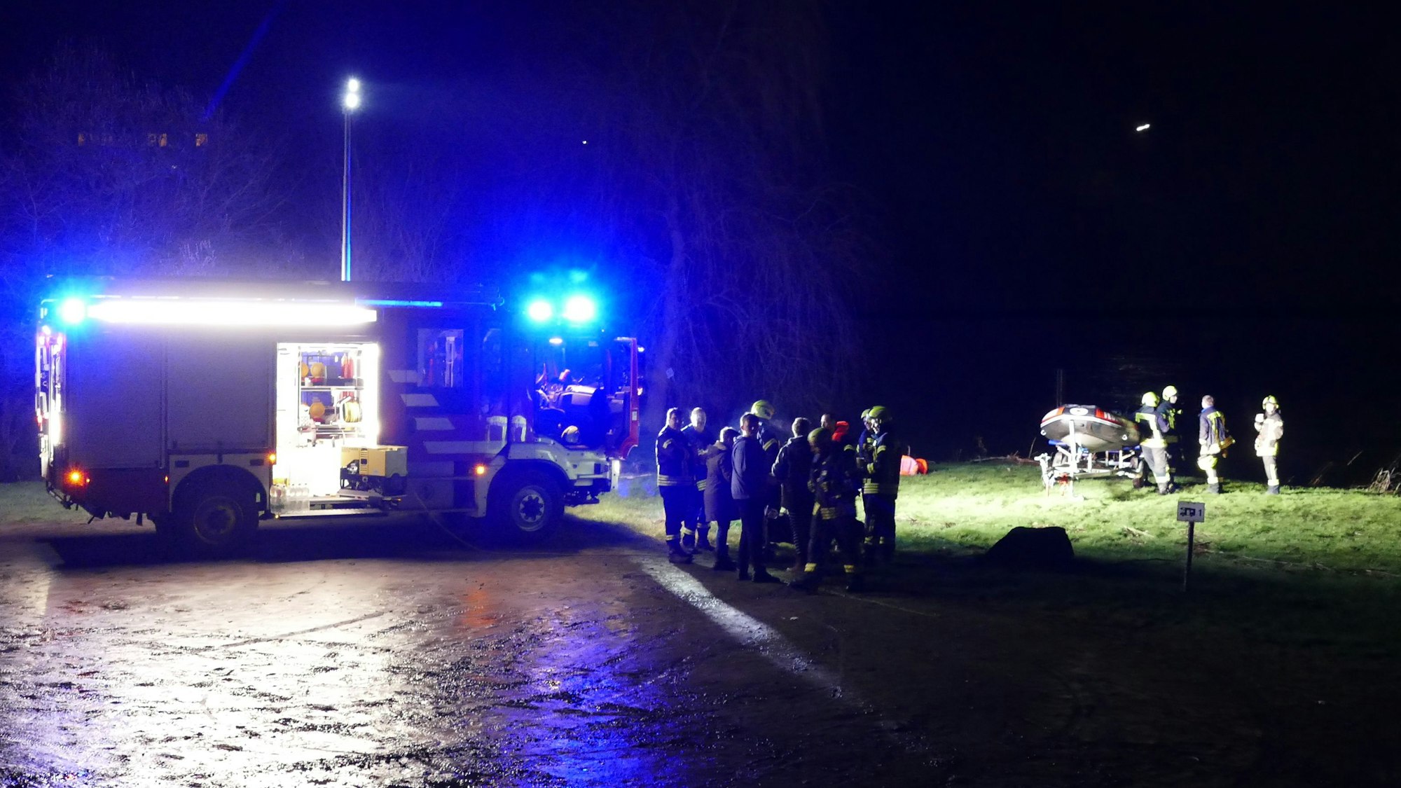 Am nächtlichen Siegufer stehen ein Feuerwehrauto und Feuerwehrleute, auch ein Schlauchboot ist am Ufer.