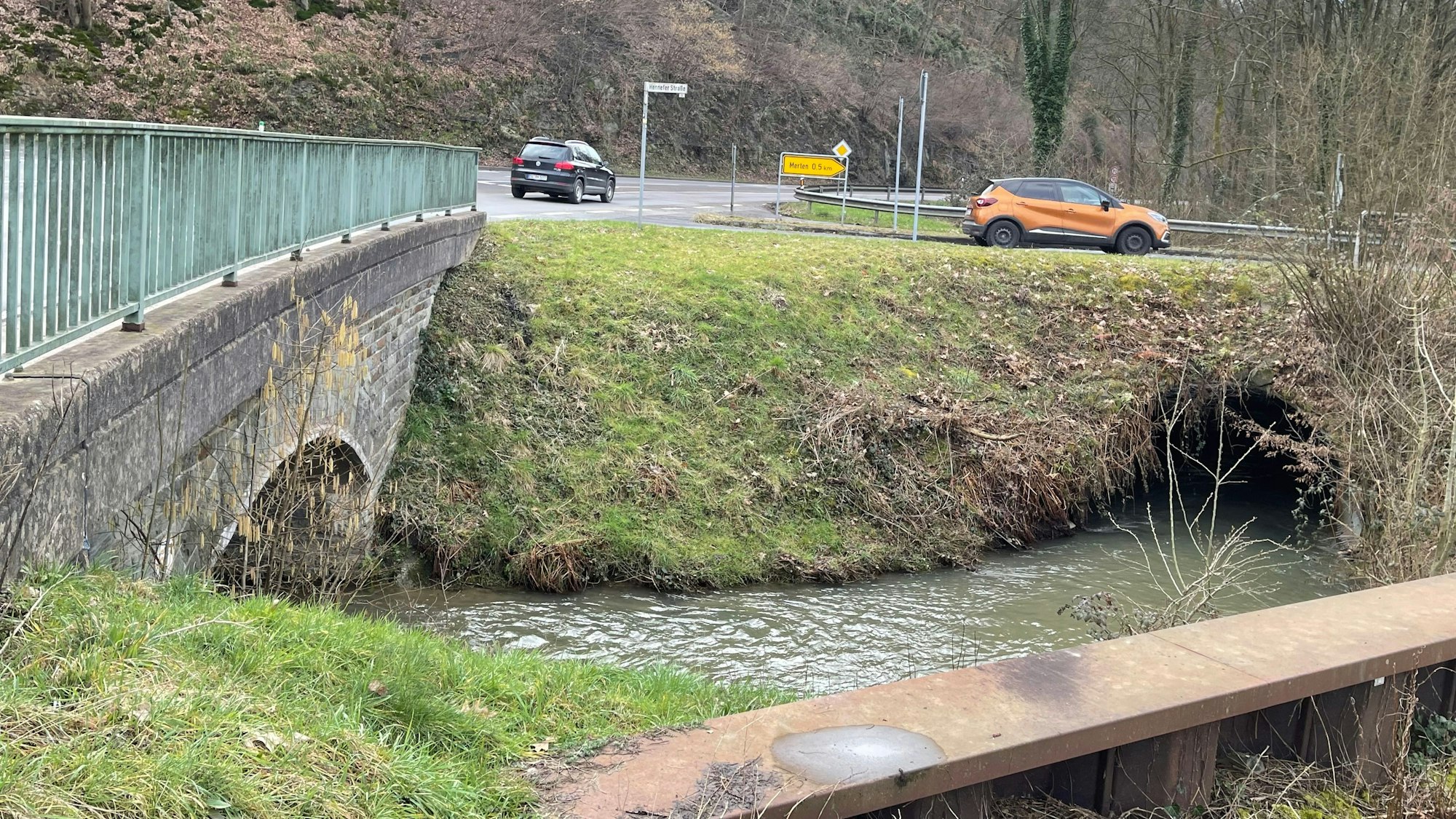 Ein breiter Bach fließt durch zwei große Tunnel, vom Vordergrund ist eine Wand aus rostigem Wellstahl. Ein Wegweiser zeigt nach Merten, zwei Autos fahren auf der Straße hinter der Brücke.