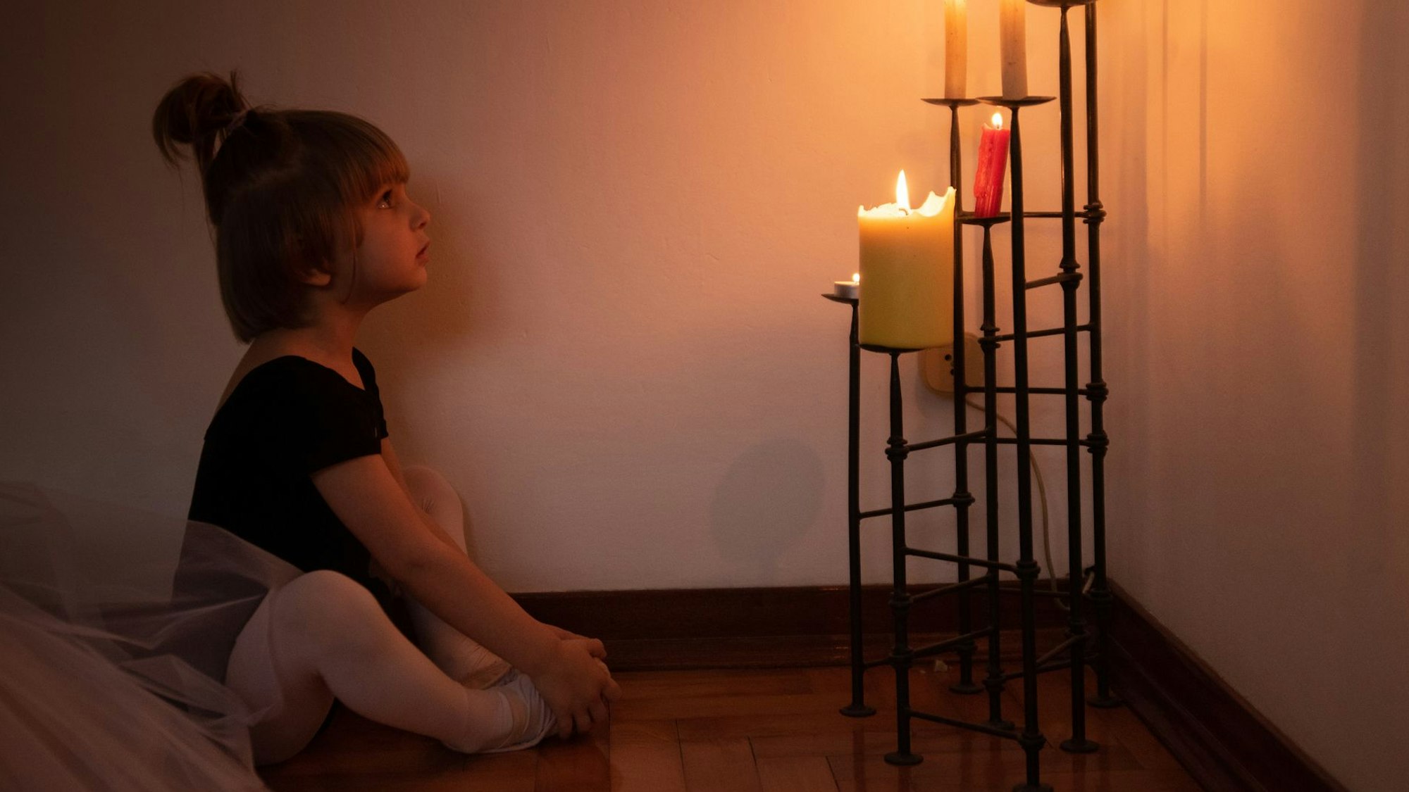 Ein kleines Mädchen sitzt im Schneidersitz auf dem Boden und betrachtet einen Kerzenständer mit brennenden Kerzen.