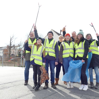 Eine Gruppe Frauen, Männer und Kinder in gelben Warnwesten und mit Mülltüten schwenken ihre Greifzangen.