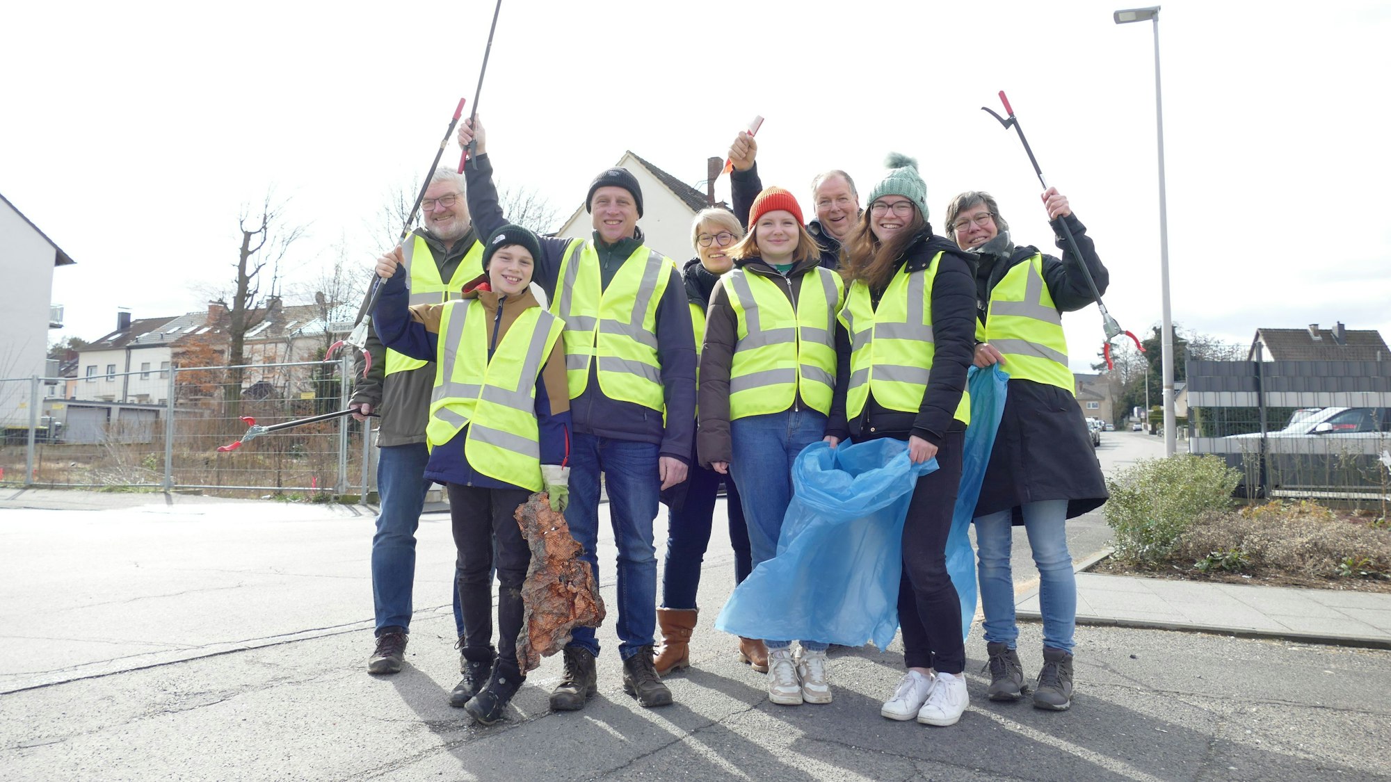 Eine Gruppe Frauen, Männer und Kinder in gelben Warnwesten und mit Mülltüten schwenken ihre  Greifzangen.