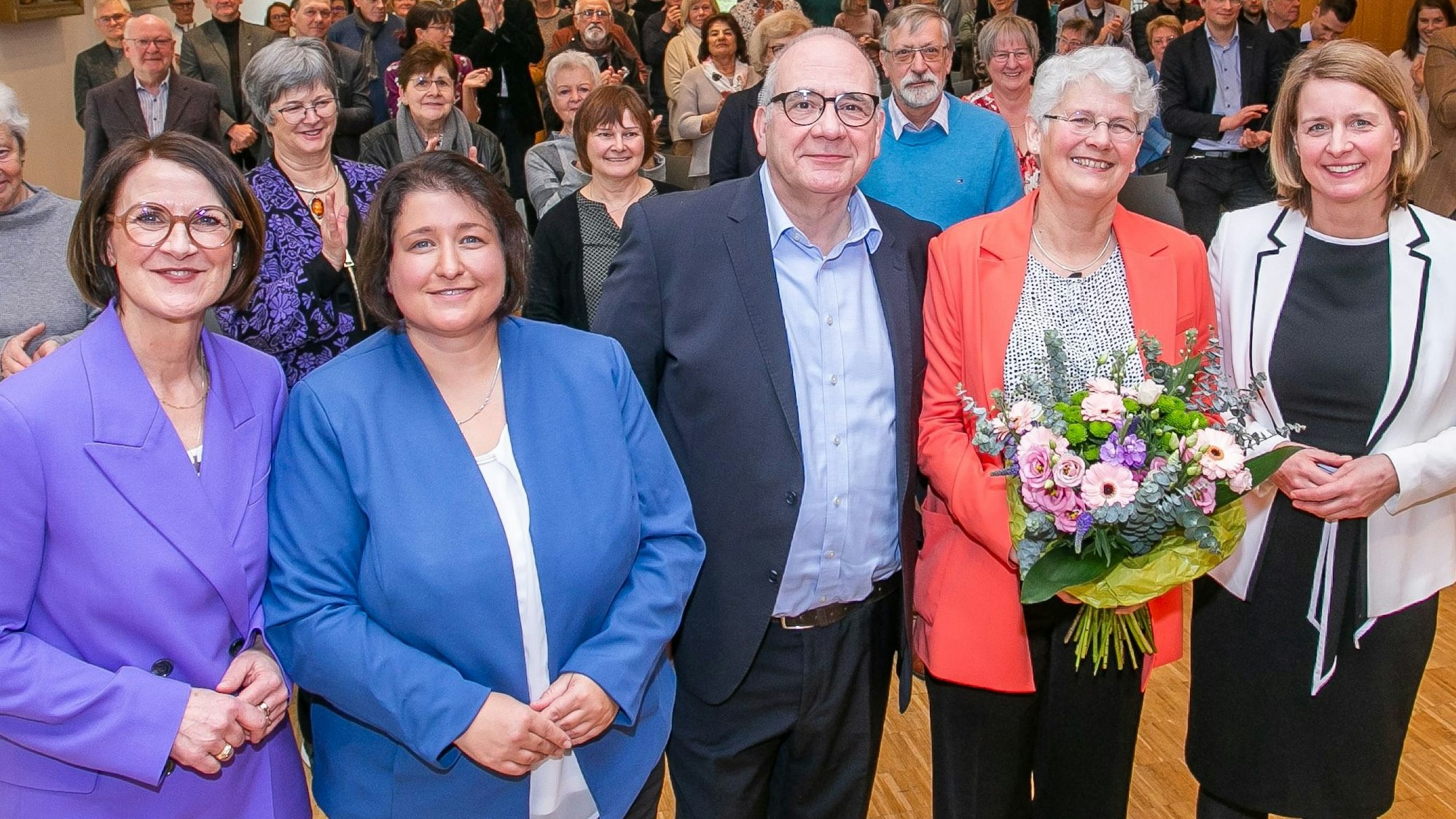 Bärbel Schröder, zweite von rechts, steht mit einem Blumenstrauß in der Hand zwischen Gratulierenden und lächelt in die Kamera.