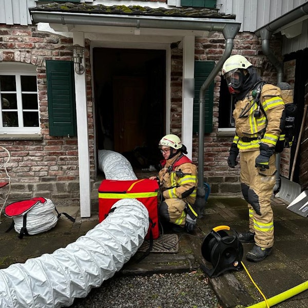 Zwei Feuerwehrleute in Schutzausrüstung installieren ein großes Lüftungsgebiet mit Schläuchen, die in ein Haus hineinführen.