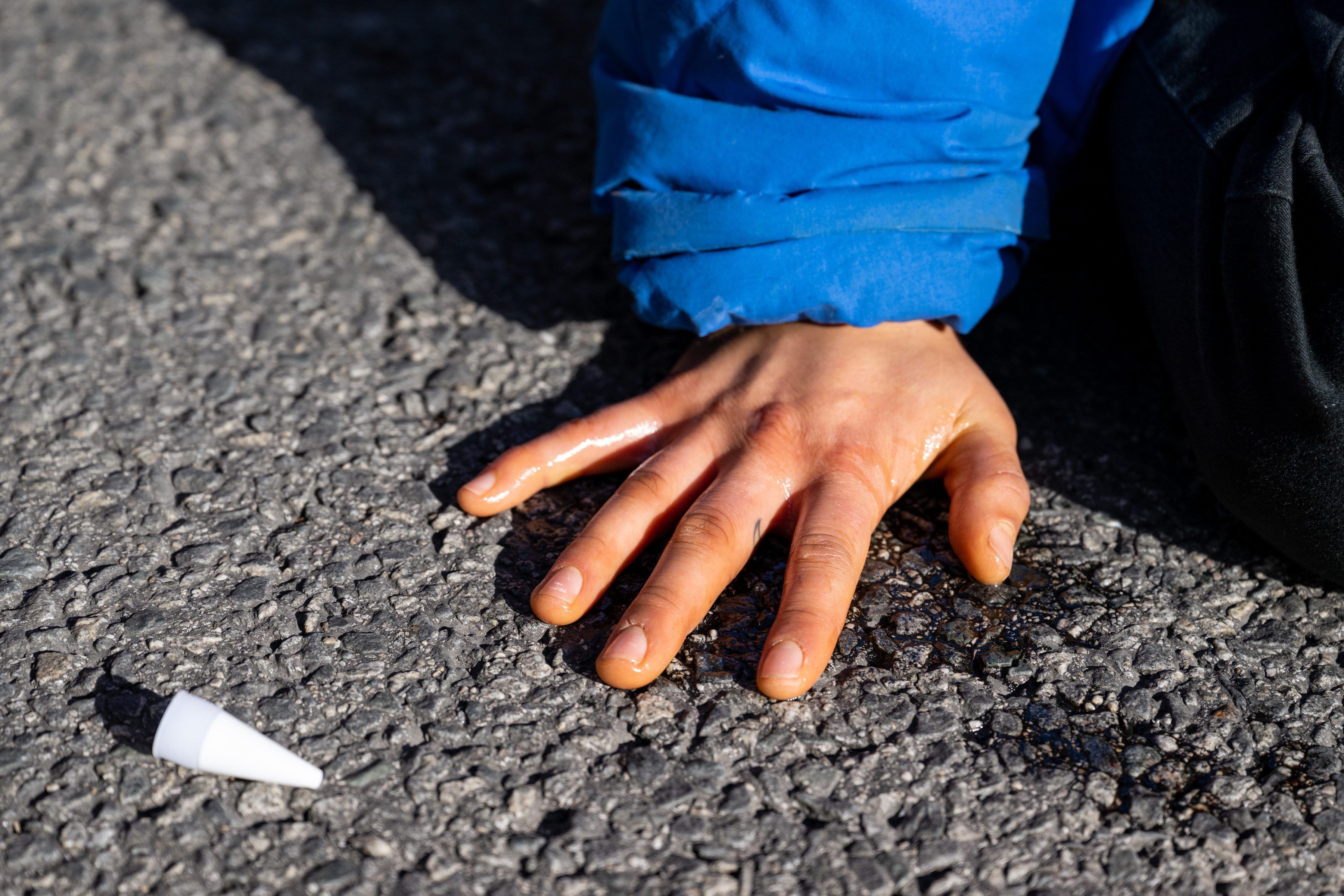 Eine Hand ist festgeklebt auf Asphalt. Daneben liegt ein kleines Plastikgefäß.