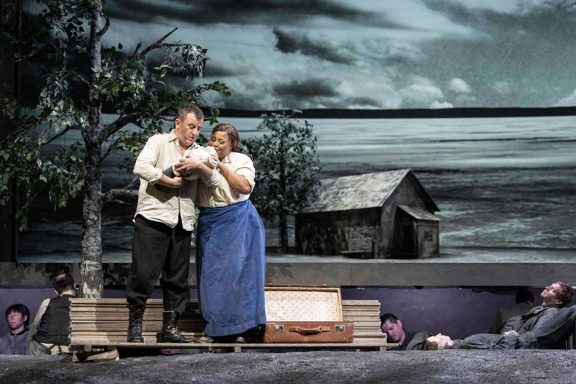 Vassili hält ein Baby in den Händen, Stephana ist dicht neben ihm. Hinter den beiden wächst ein Baum. Im Hintergrund liegen Schauspieler des Ensembles wie leblos auf dem Boden.