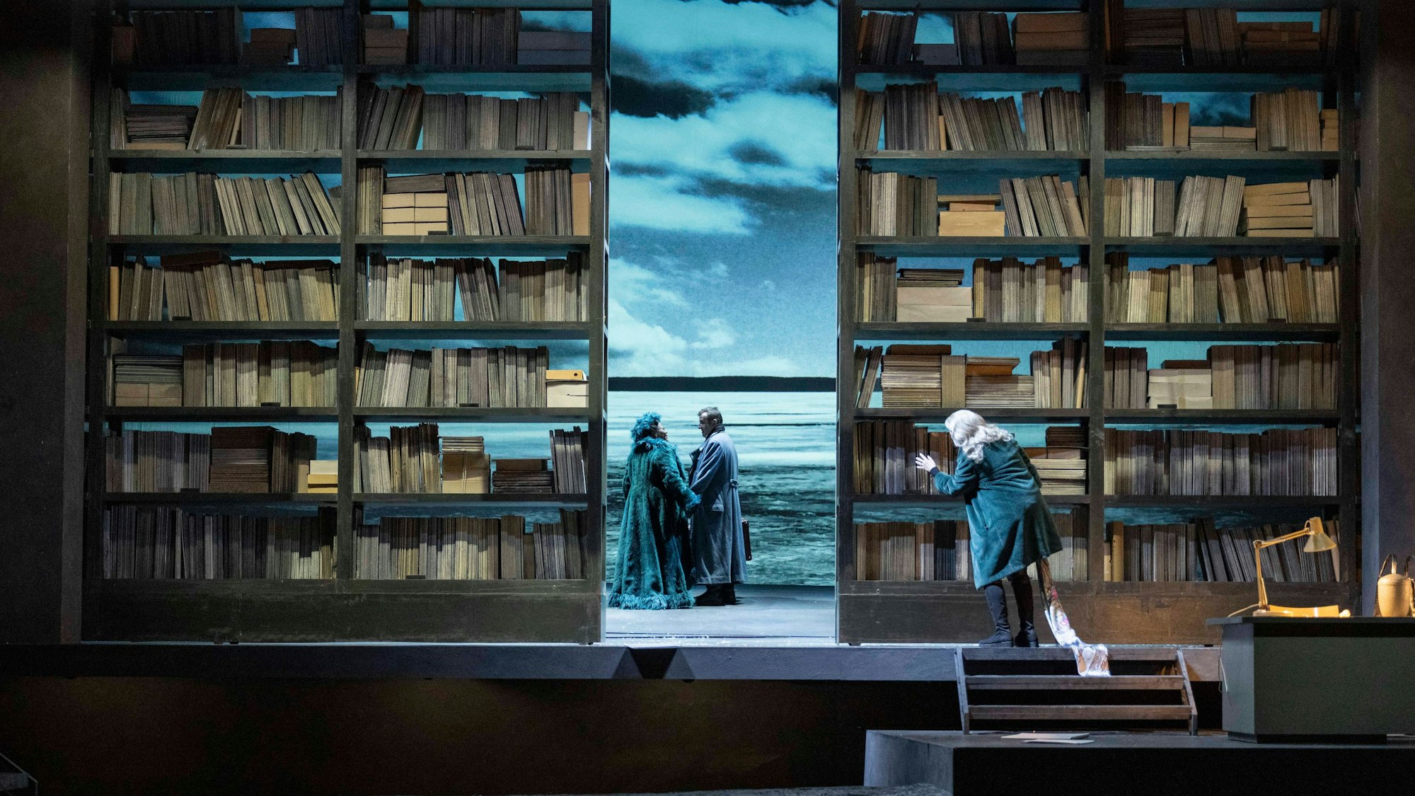 Auf der Bühne stehen zwei sehr hohe Bücherschränke, in der Mitte zeigt ein Spalt einen Strand, wo ein Paar sich an den Händen hält.