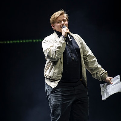 Sebastian Hotz steht in einem blauen Pullover und einer hellen Jacke auf einer Bühne. Er spricht in ein Mikrofon