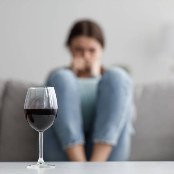 Sad caucasian millennial female sits on sofa suffering from stress and troubles, focus on bottle and glass of wine in home interior.