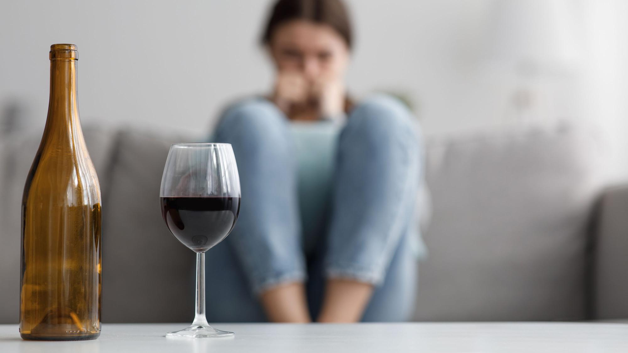 Sad caucasian millennial female sits on sofa suffering from stress and troubles, focus on bottle and glass of wine in home interior.