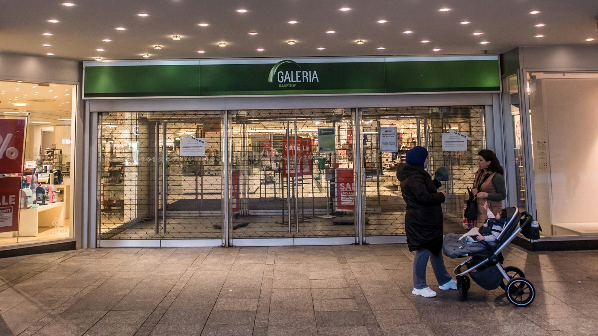 Zwei Frauen stehen mit einem Kinderwagen vor der geschlossenen Tür des Galeria-Warenhauses in Leverkusen.