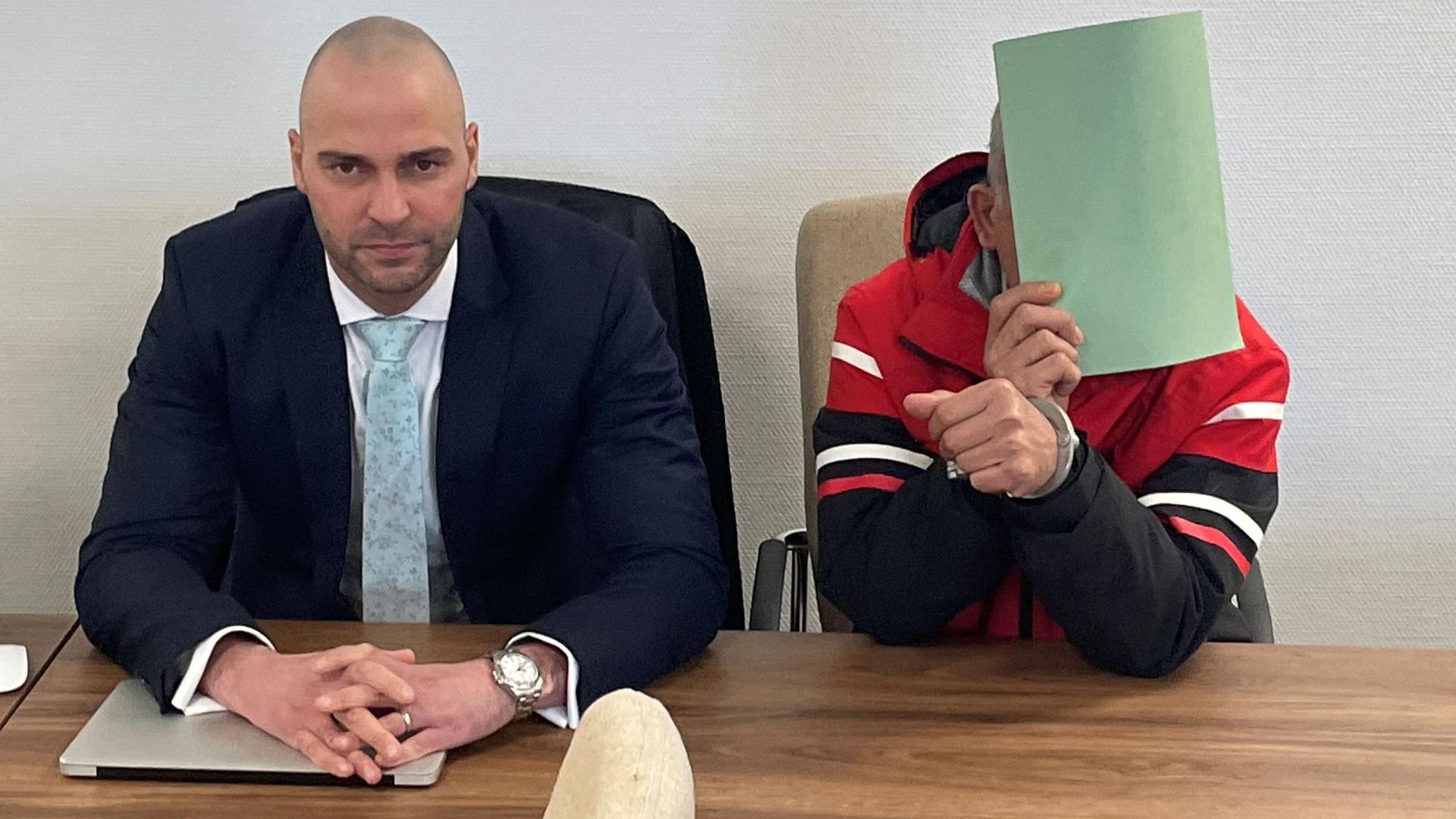 Der Angeklagte mit seinem Verteidiger Jan Victor Khatib beim Prozessauftakt im Kölner Landgericht. Der Angeklagte hält sich eine Kladde vors Gesicht.