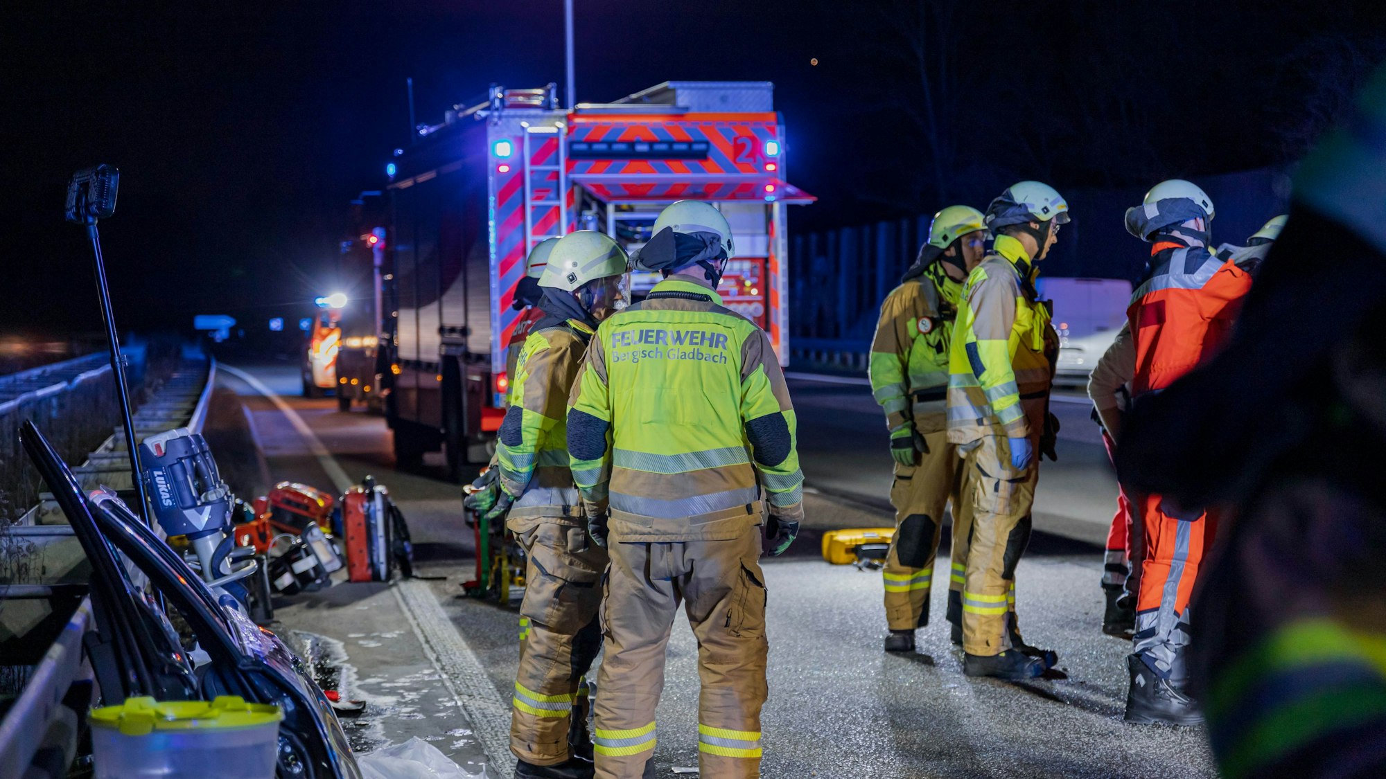 Feuerwehrleute stehen an einem Einsatzfahrzeug mit Blaulicht auf der Autobahn. An der Leitplanke lehnen abgetrennte Autoteile wie zwei Autotüren.
