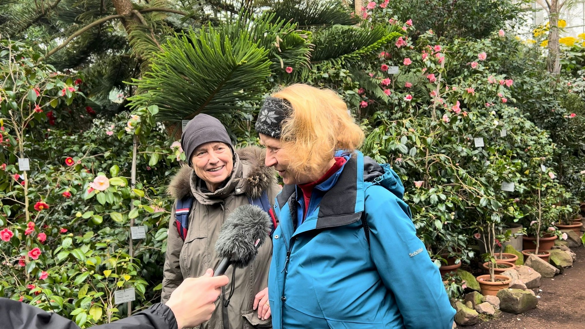 Zwei Frauen stehen vor Büschen und Blumen.