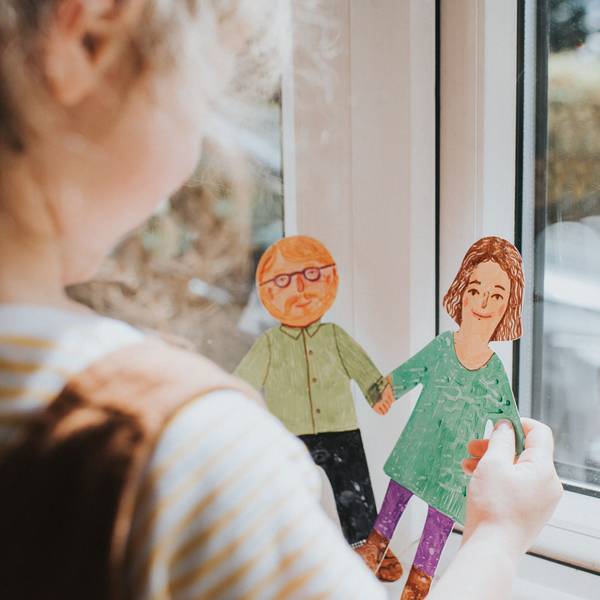 Ein kleines Mädchen steht am Fenster und hält zwei bemalte Pappfiguren in der Hand, eine Frau und einen Mann mit Bart und Brille.